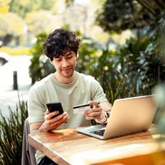 Ein junger Mann sitzt mit Handy und Kreditkarte am Laptop