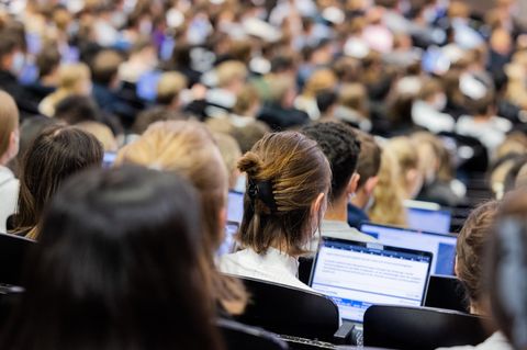 Eine Studentin sitzt im Hörsaal zwischen anderen Studenten und schaut in ihr Laptop.