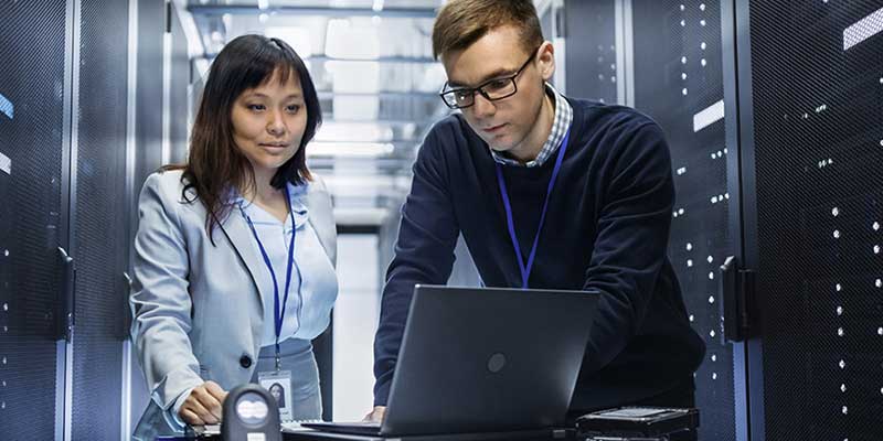 Two employees in a room of servers, looking at a laptop