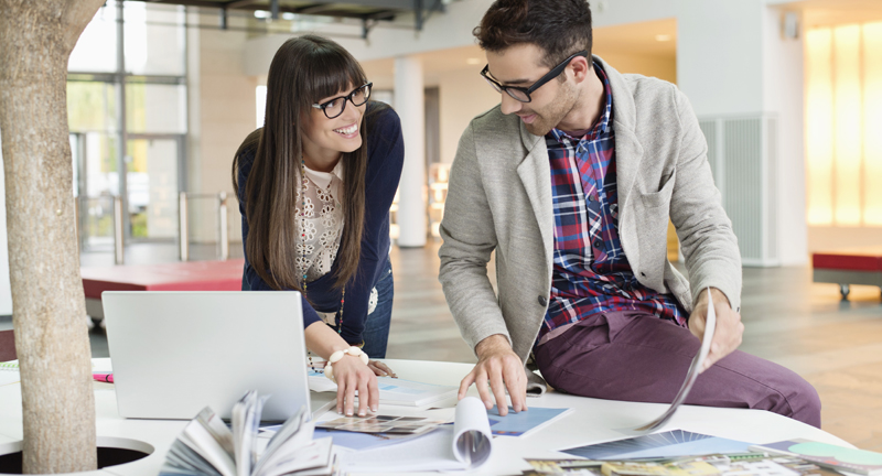 A woman and a man in a brightly lit office, sitting on a table, looking at print samples