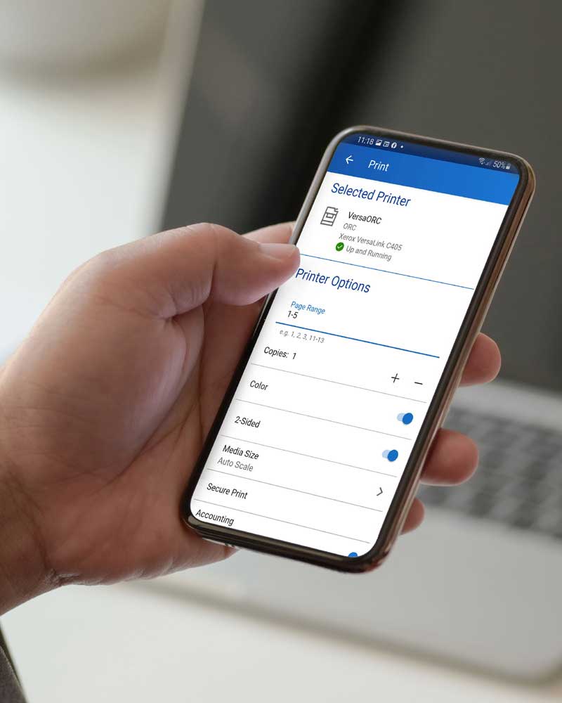 Hand holding a smartphone showing the Xerox Workplace Mobile App with a laptop in the background