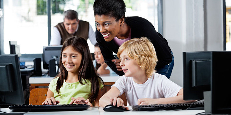 Children sitting at table with teacher working on computers