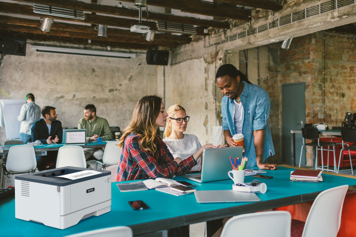 Coworkers in an office with brick walls and blue tables, using a laptop next to a Xerox B310 Printer