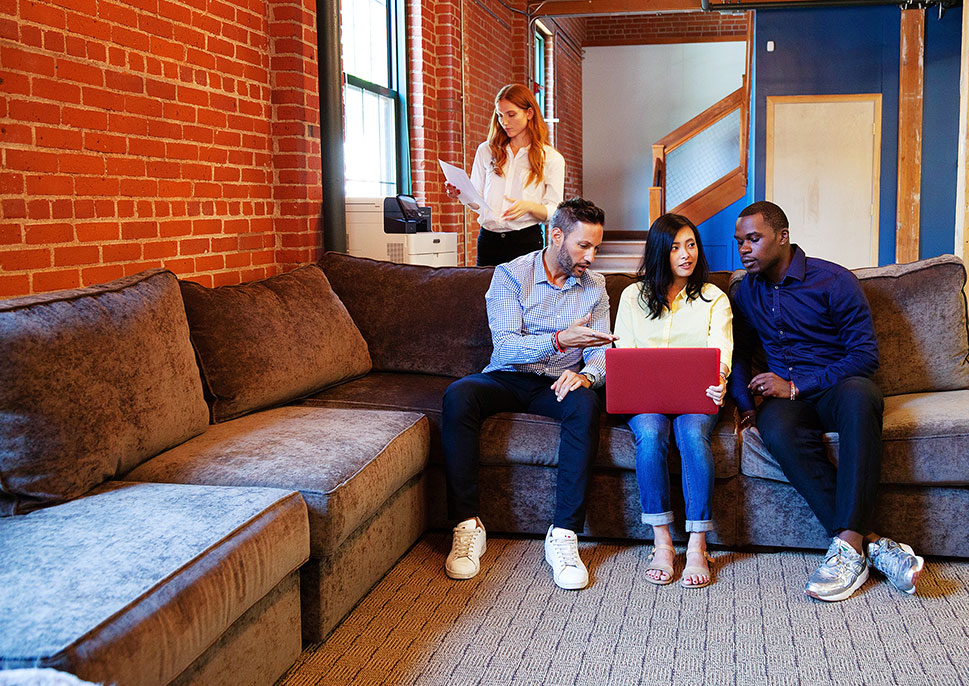 3 people sitting on a couch with a laptop. Behind them is a woman picking up prints from a Xerox WorkCentre MFP.