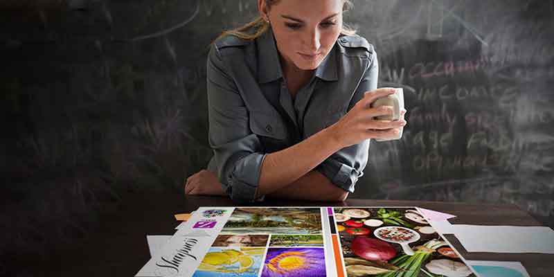 Woman with coffee, in front of a chalk board, looking at a table covered with print samples