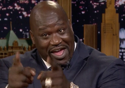 Shaquille in suit laughing and pointing towards the camera on a talk show set