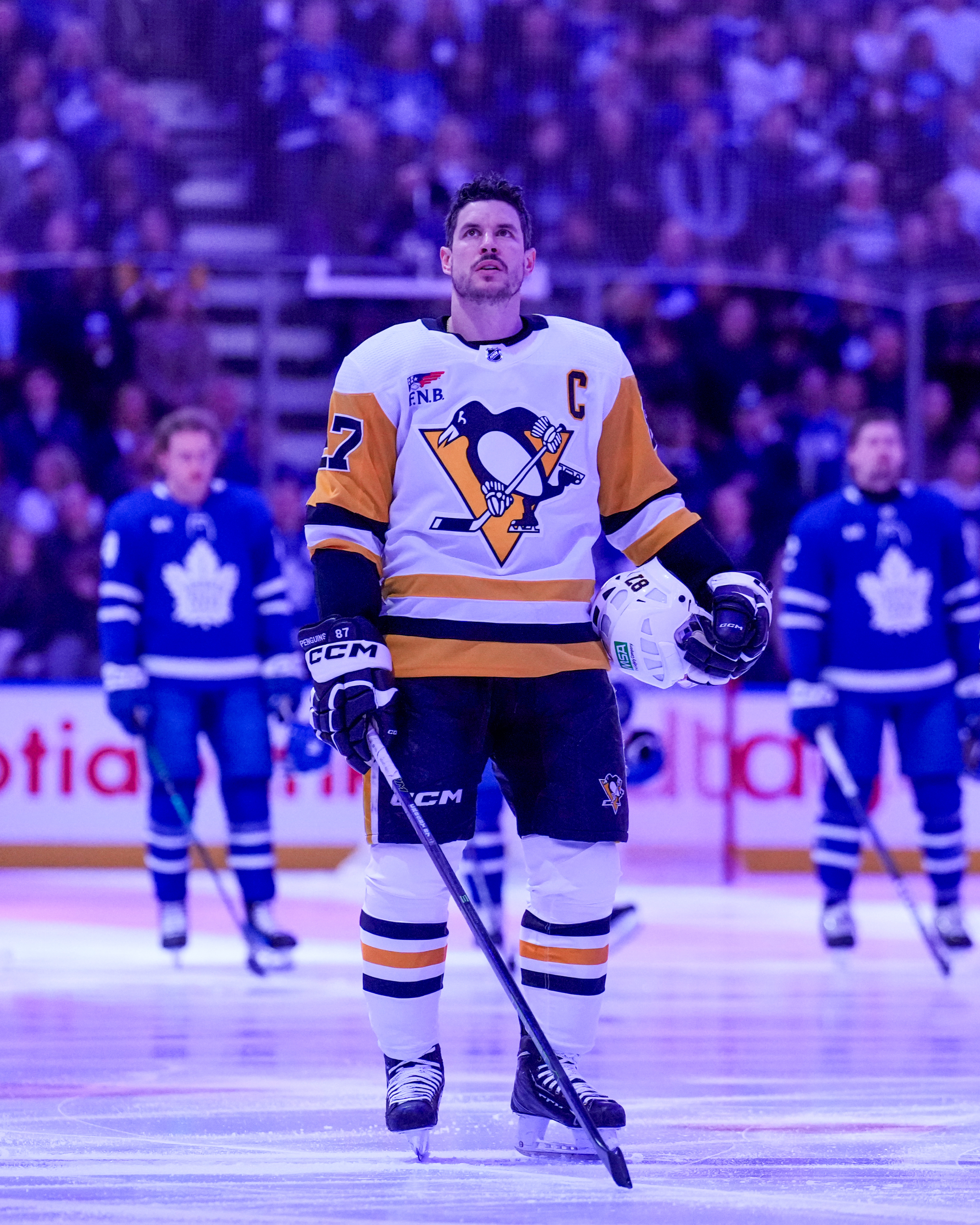 Sidney Crosby standing on ice in Pittsburgh Penguins uniform, holding a hockey stick. Toronto Maple Leafs players are in the background during the national anthem