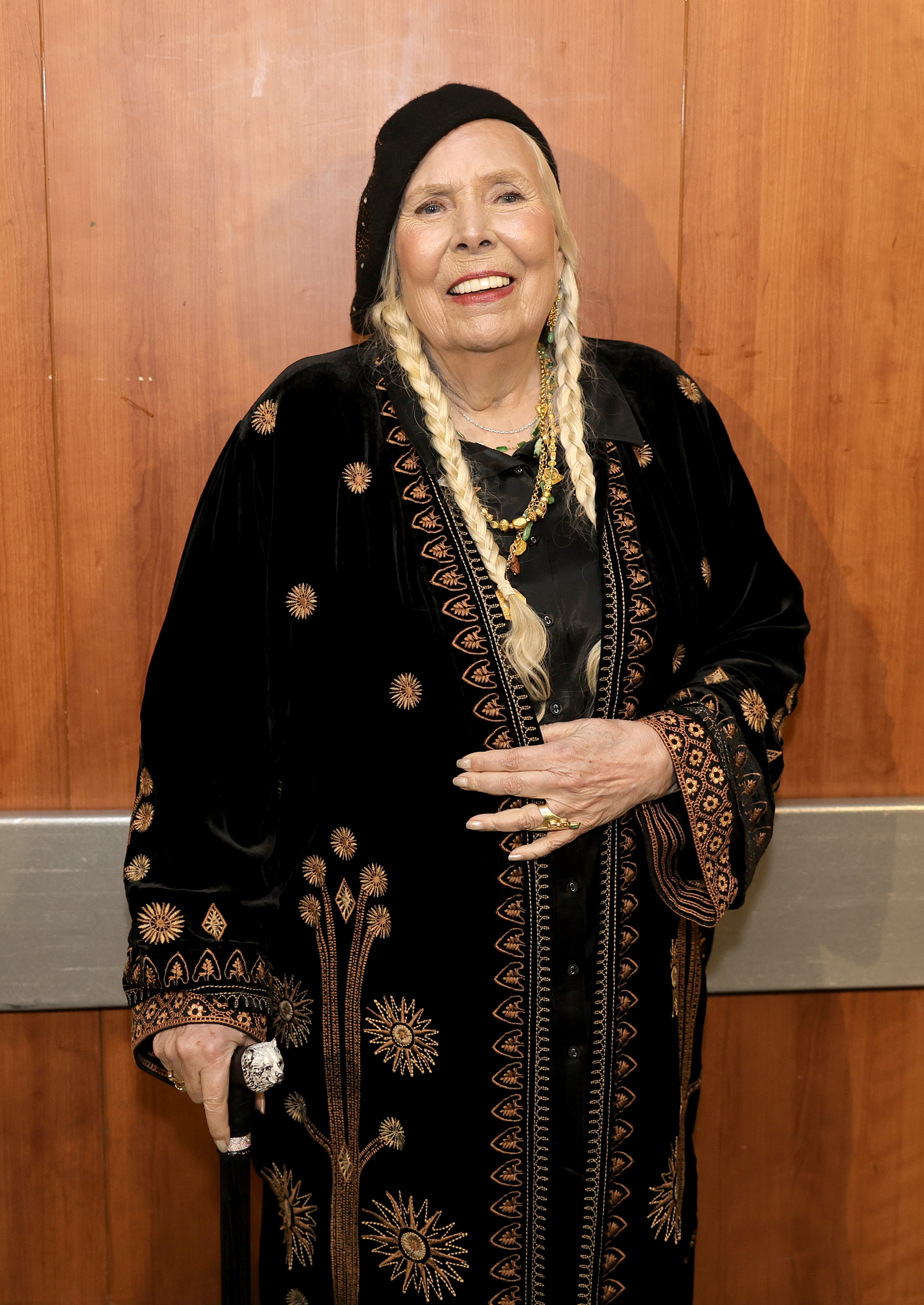 Joni Mitchell smiling, wearing a long, ornate black robe with intricate patterns and a black beret. She is holding a cane and is standing against a wooden background