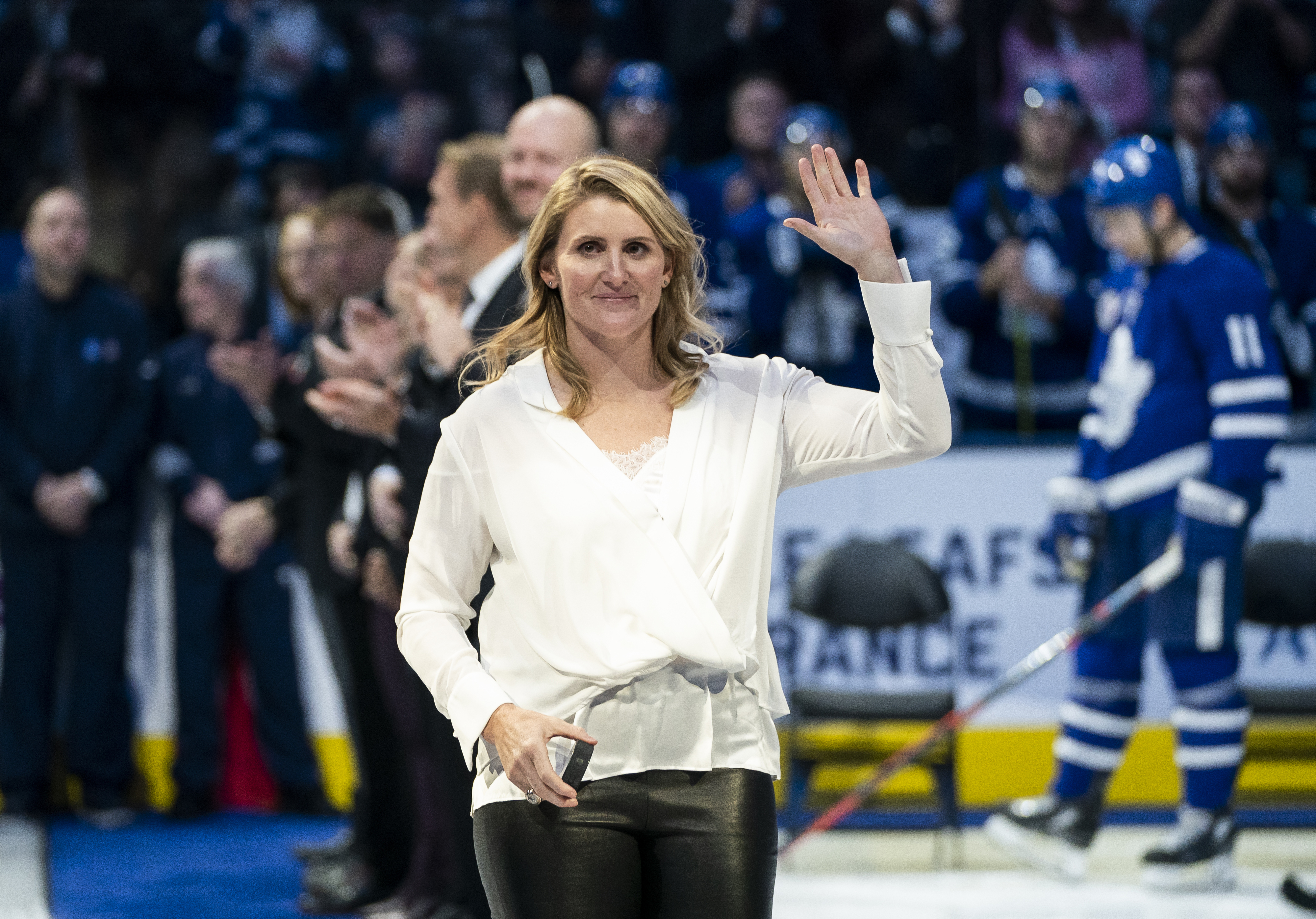 A woman in a white blouse and black pants, holding a phone, waves to the crowd at a hockey event. Hockey players and audience members are in the background