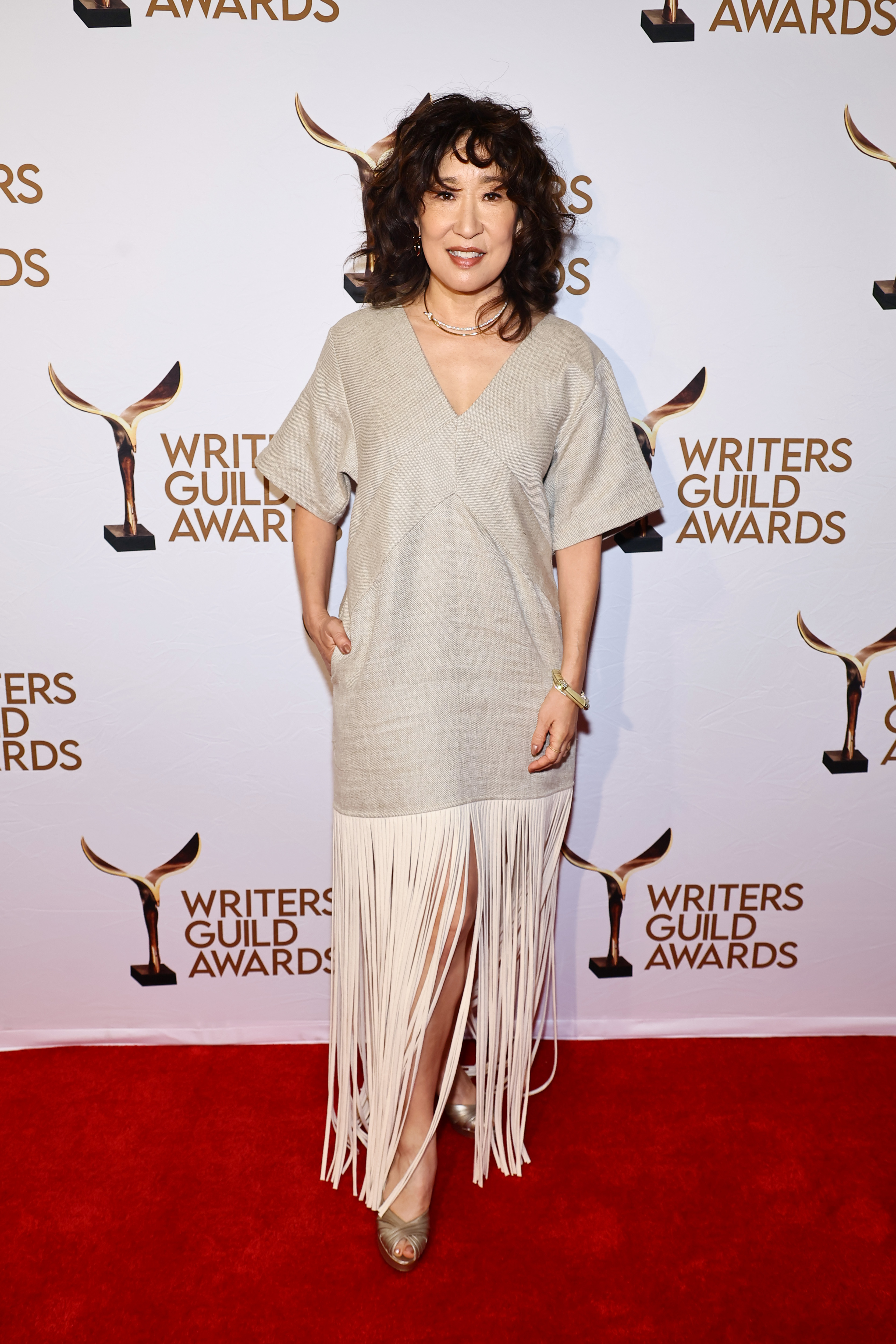 Sandra Oh stands on the red carpet at the Writers Guild Awards, wearing a light-colored dress with fringe detailing at the bottom