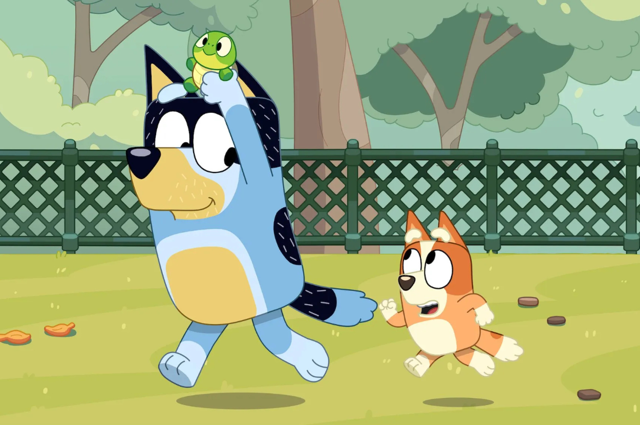 Bluey, holding a green toy frog, skips cheerfully while Bingo runs behind in a park setting with trees and a fence