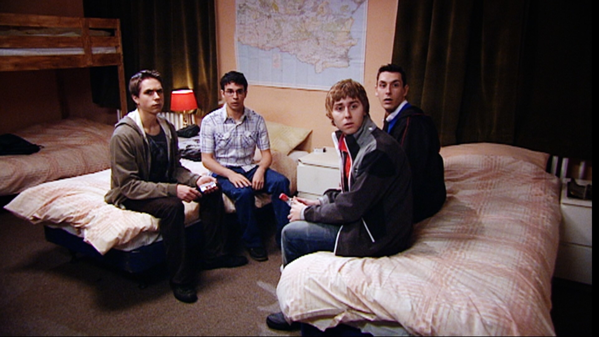 Simon Bird, Joe Thomas, James Buckley, and Blake Harrison in a bedroom scene from The Inbetweeners, all sitting on beds looking towards the camera with surprised expressions