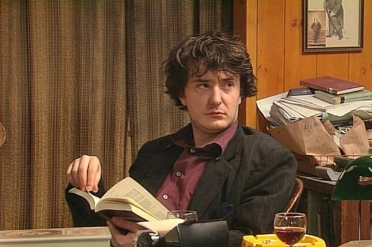 Dylan Moran in character as Bernard Black from the TV show &quot;Black Books,&quot; seated at a cluttered desk holding an open book and looking to the side