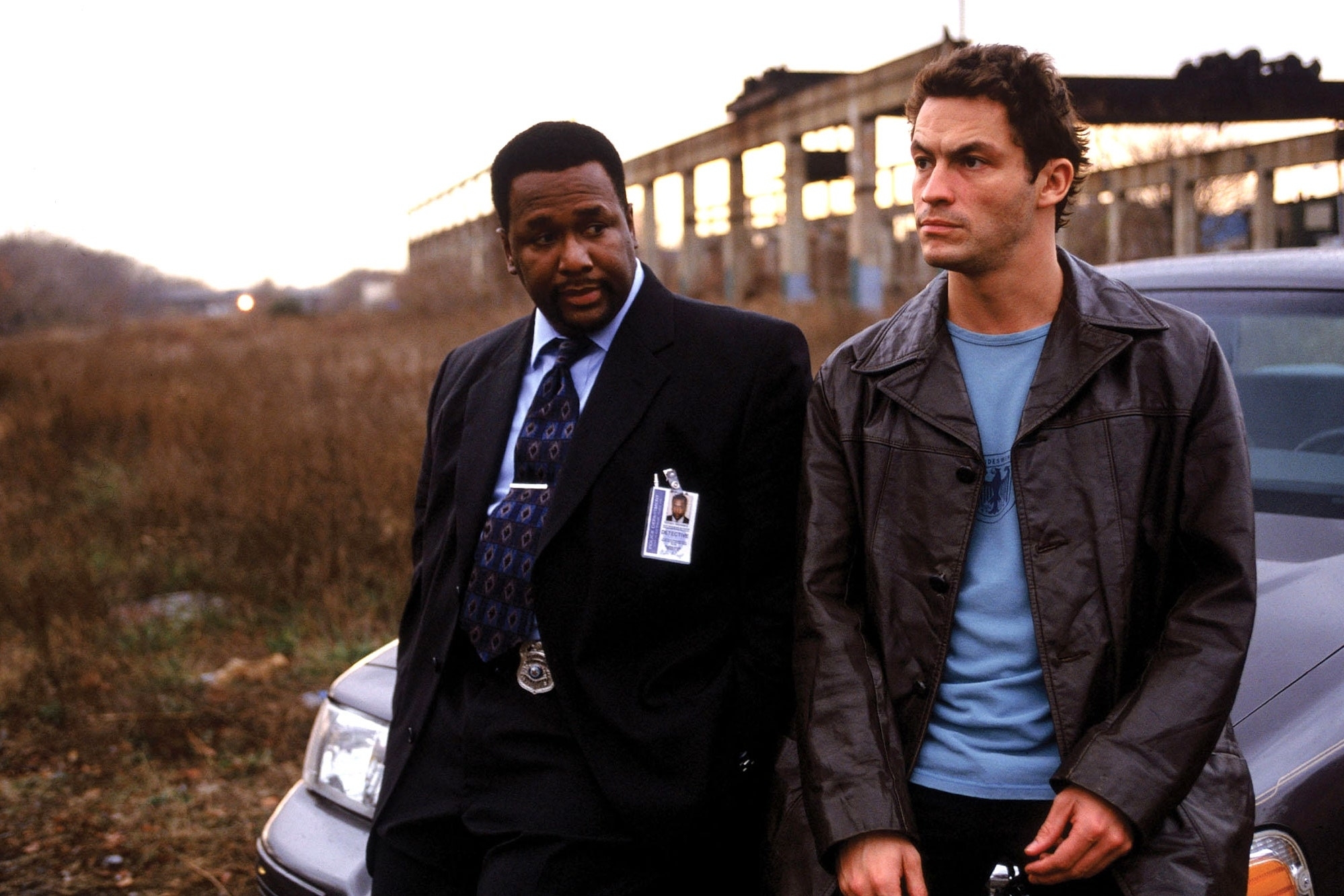 Wendell Pierce in a suit and badge, and Dominic West in a casual jacket, lean against a car with an industrial backdrop from &quot;The Wire.&quot;