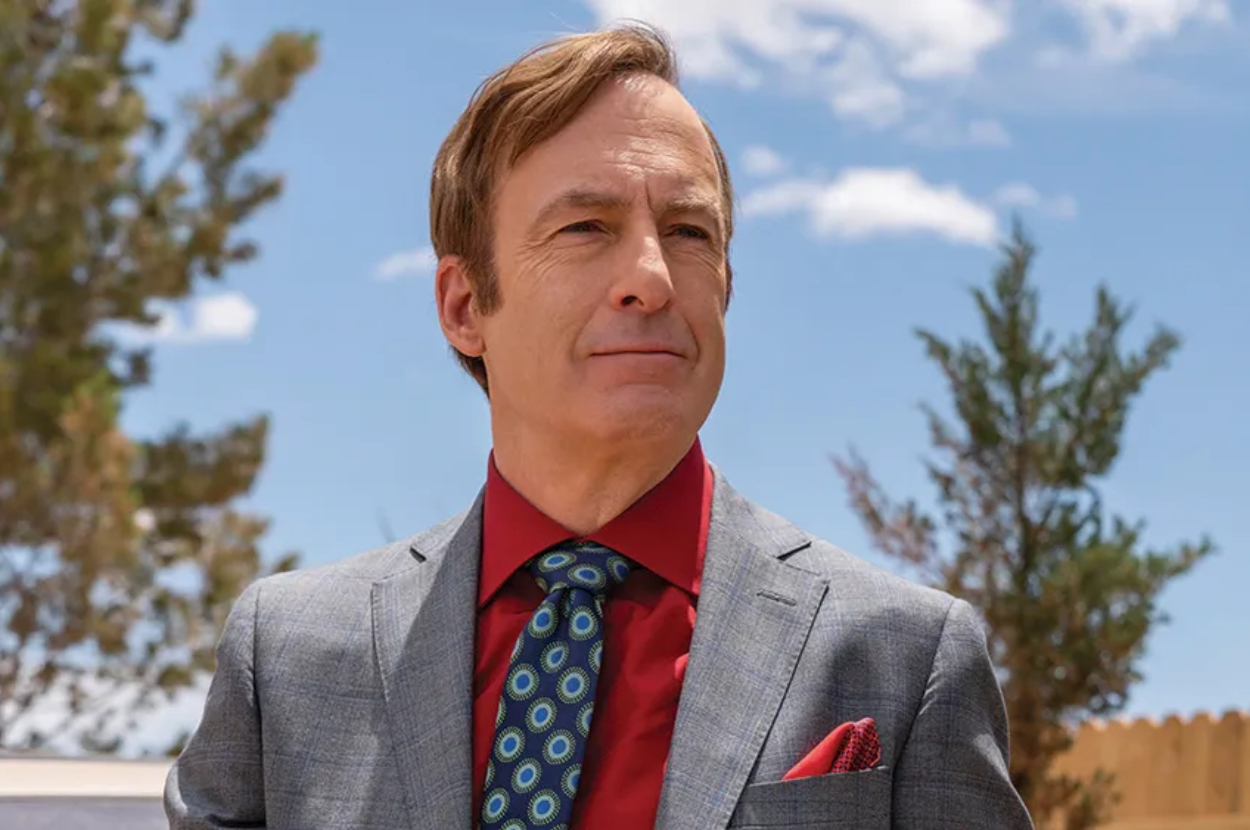 Bob Odenkirk dressed in a grey suit with a patterned tie and red shirt, outdoors with trees and sky in the background