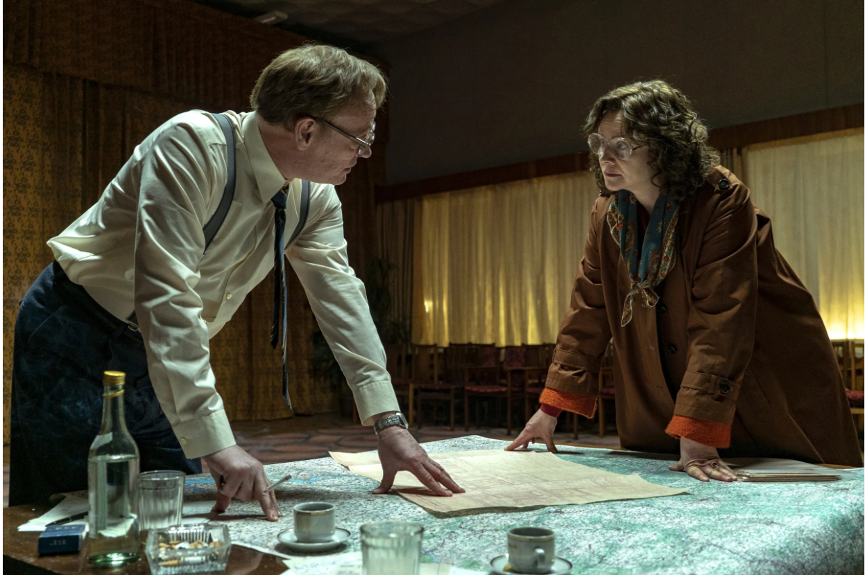 Jared Harris and Emily Watson in a scene from Chernobyl, leaning over a table with a map, discussing seriously. Vintage setting with coffee cups