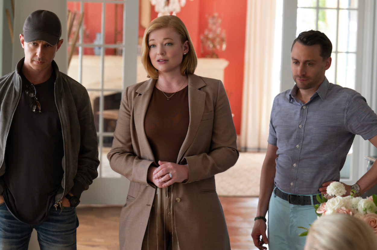 Jeremy Strong, Sarah Snook, and Kieran Culkin stand indoors, with Sarah in a blazer, Jeremy in a casual jacket and cap, and Kieran in a buttoned shirt