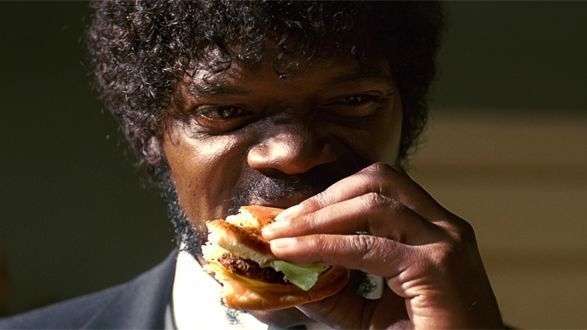 A man with curly hair and a beard, wearing a suit, takes a bite of a burger