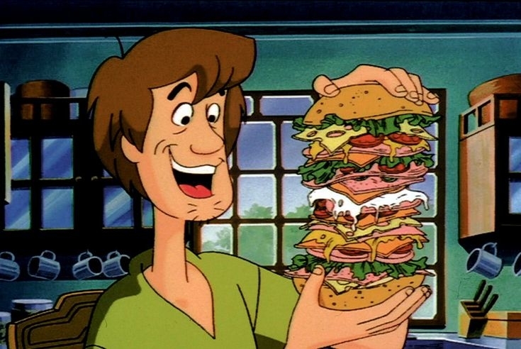 Shaggy from Scooby-Doo is holding a tall, multi-layered sandwich with a delighted expression in a kitchen setting