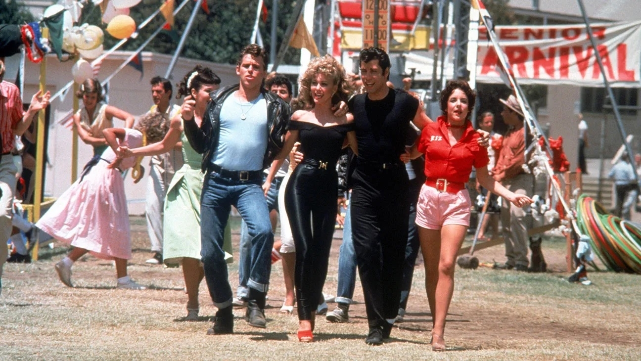 John Travolta and Olivia Newton-John walk with cast members at a fairground in a scene from the movie Grease