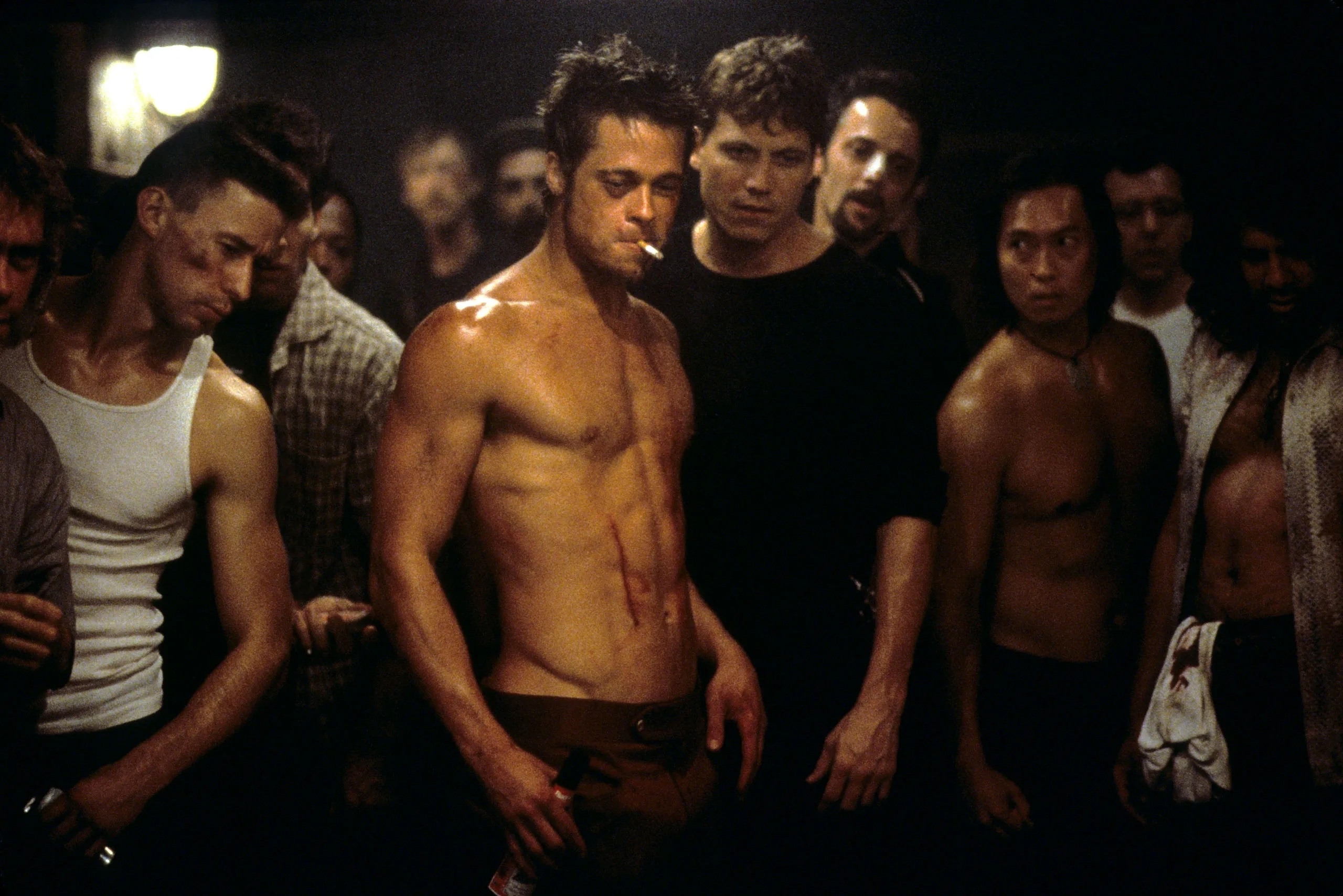 Brad Pitt, shirtless, stands in the center of a fight club with other shirtless and clothed men looking on in the background