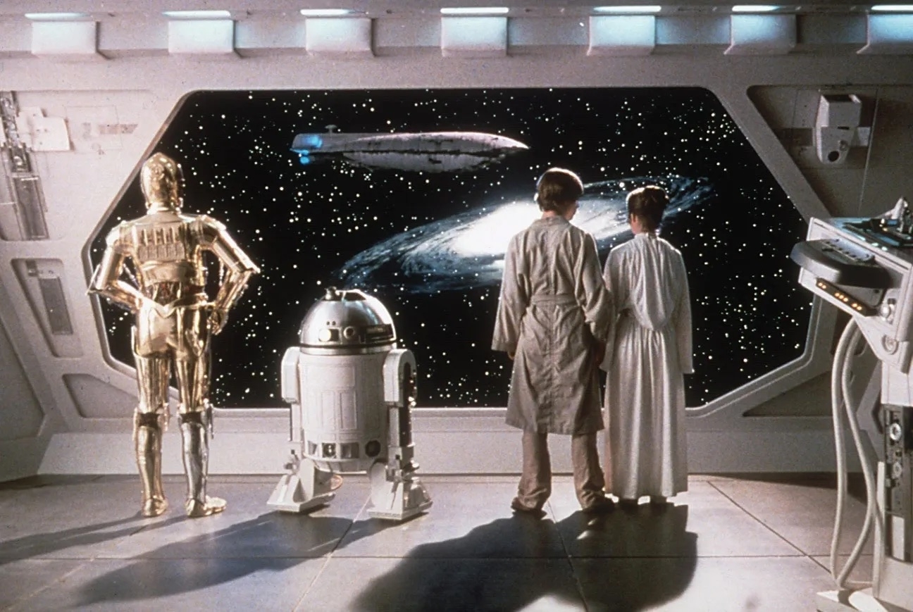 C-3PO, R2-D2, Luke Skywalker, and Princess Leia from Star Wars stand in a spaceship looking out at a galaxy and spaceship through a large window