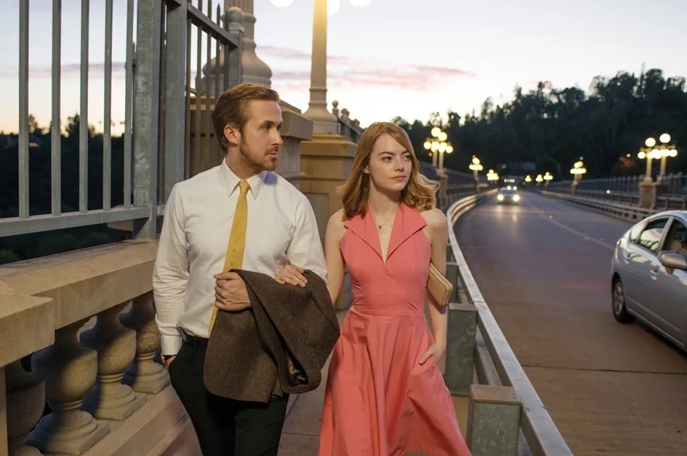 Ryan Gosling and Emma Stone walk on a bridge at sunset in a scene from &quot;La La Land.&quot; He wears a white shirt and tie, and she is in a pink dress