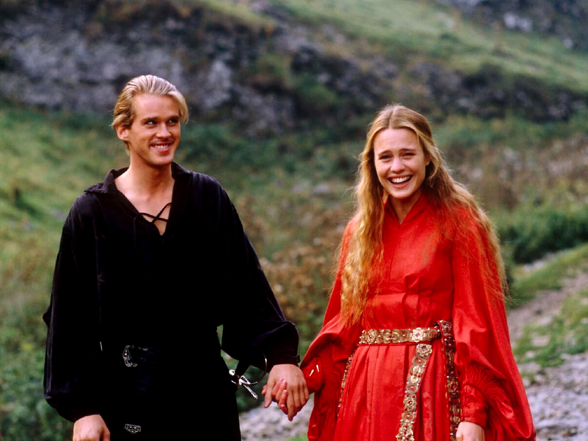 Cary Elwes, in black medieval attire, holds hands with Robin Wright, in a red medieval gown, as they walk through a scenic outdoor setting from &quot;The Princess Bride.&quot;