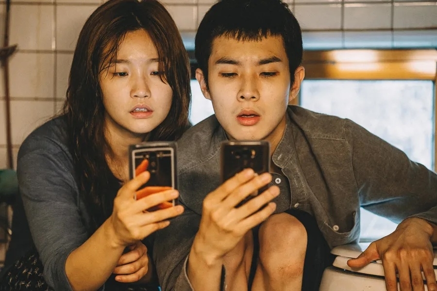 Choi Woo-shik and Park So-dam look at smartphones together in a scene from the movie &quot;Parasite.&quot; They appear focused and are sitting close to each other