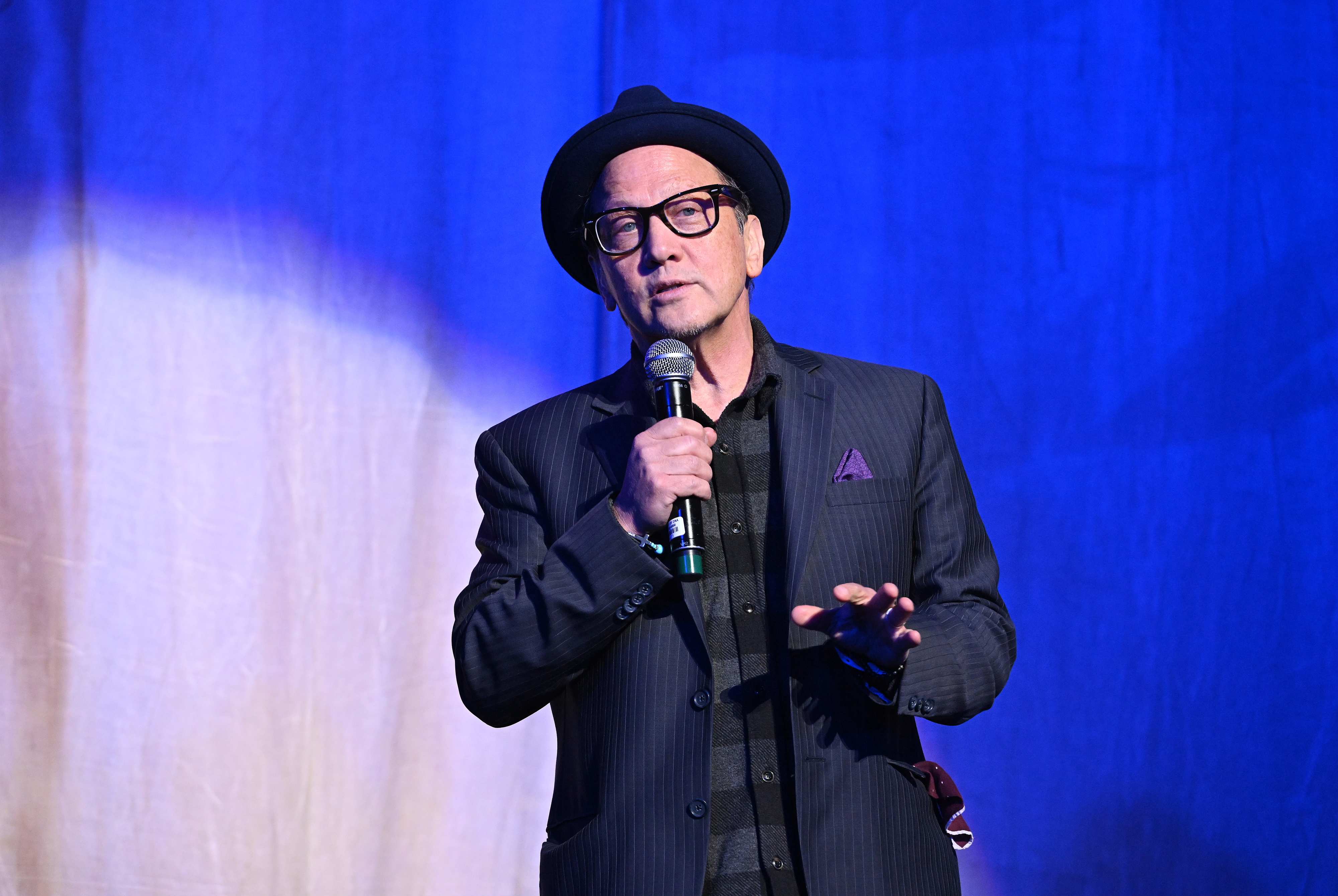 Rob Schneider wearing a hat and striped suit speaks into a microphone on stage