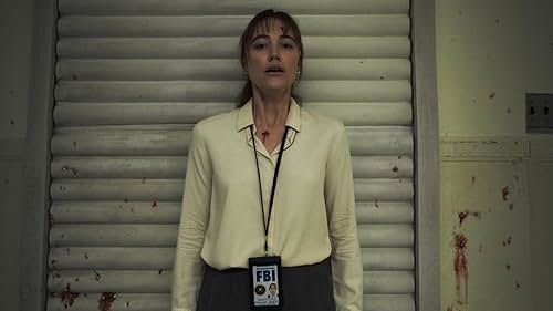 A woman stands against a metal door, wearing a light blouse and dark pants with an FBI badge around her neck