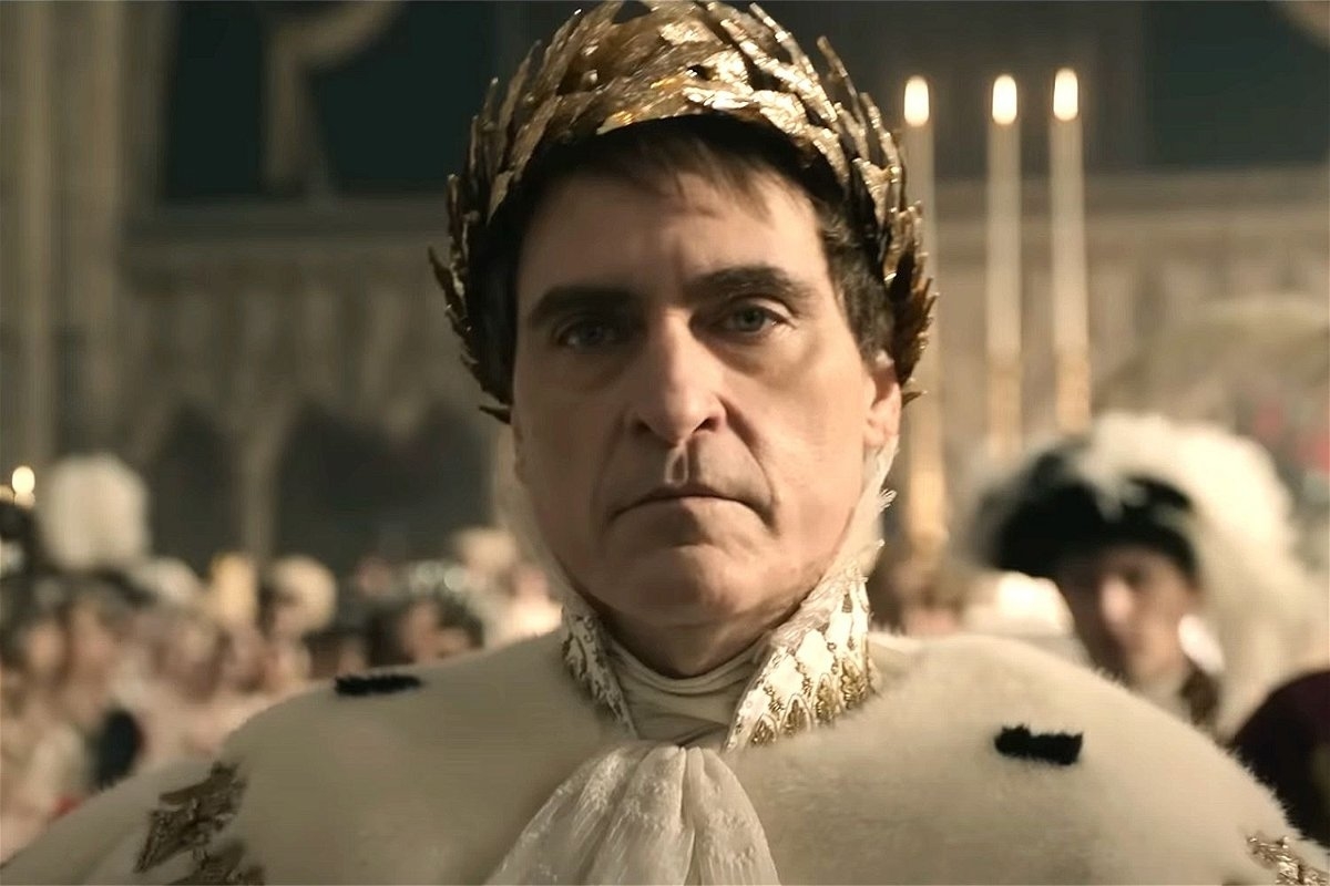 A person wearing a regal outfit and a golden laurel crown stands in an ornate setting that resembles a formal gathering or ceremony