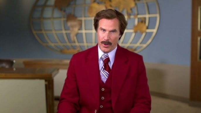 A man in a suit resembling a news anchor is seated in an office setting with a world map on the wall behind him