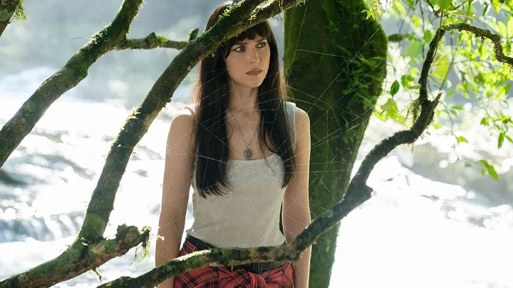 I don’t know who the person is, but they are outdoors, wearing a white top and a red plaid shirt tied at the waist, standing near tree branches