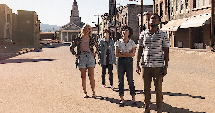 Four people stand on a deserted street. Three have casual attire, and one wears a jacket and shorts. A church is visible in the background