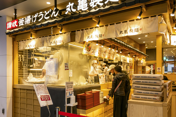 うどん専門店「丸亀製麺」の店頭の様子。顧客がうどんを注文する様子。スタッフが厨房でうどんを調理している。