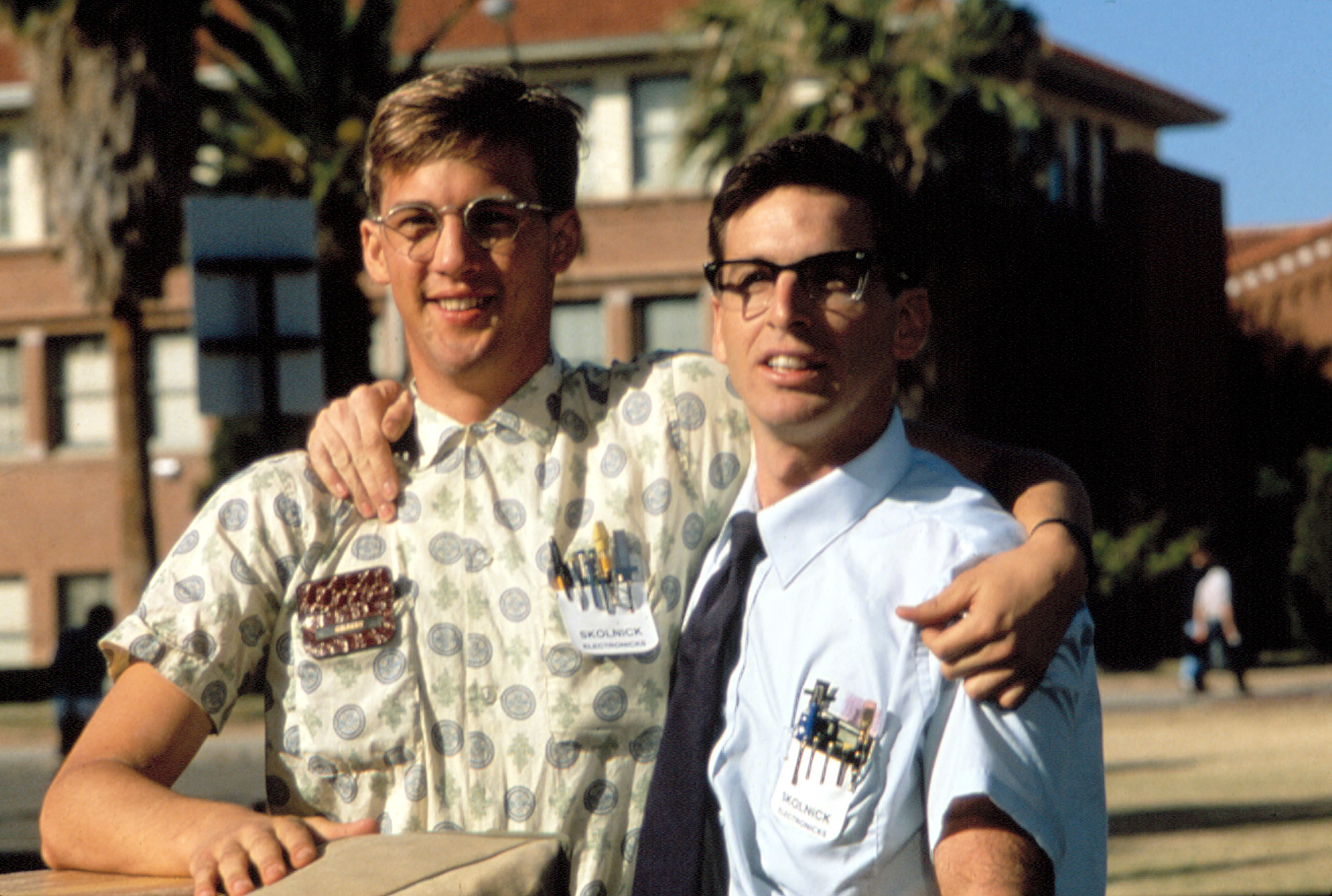 Two people outdoors, one wearing a patterned shirt, the other in a white shirt and tie, both with glasses, embracing and smiling