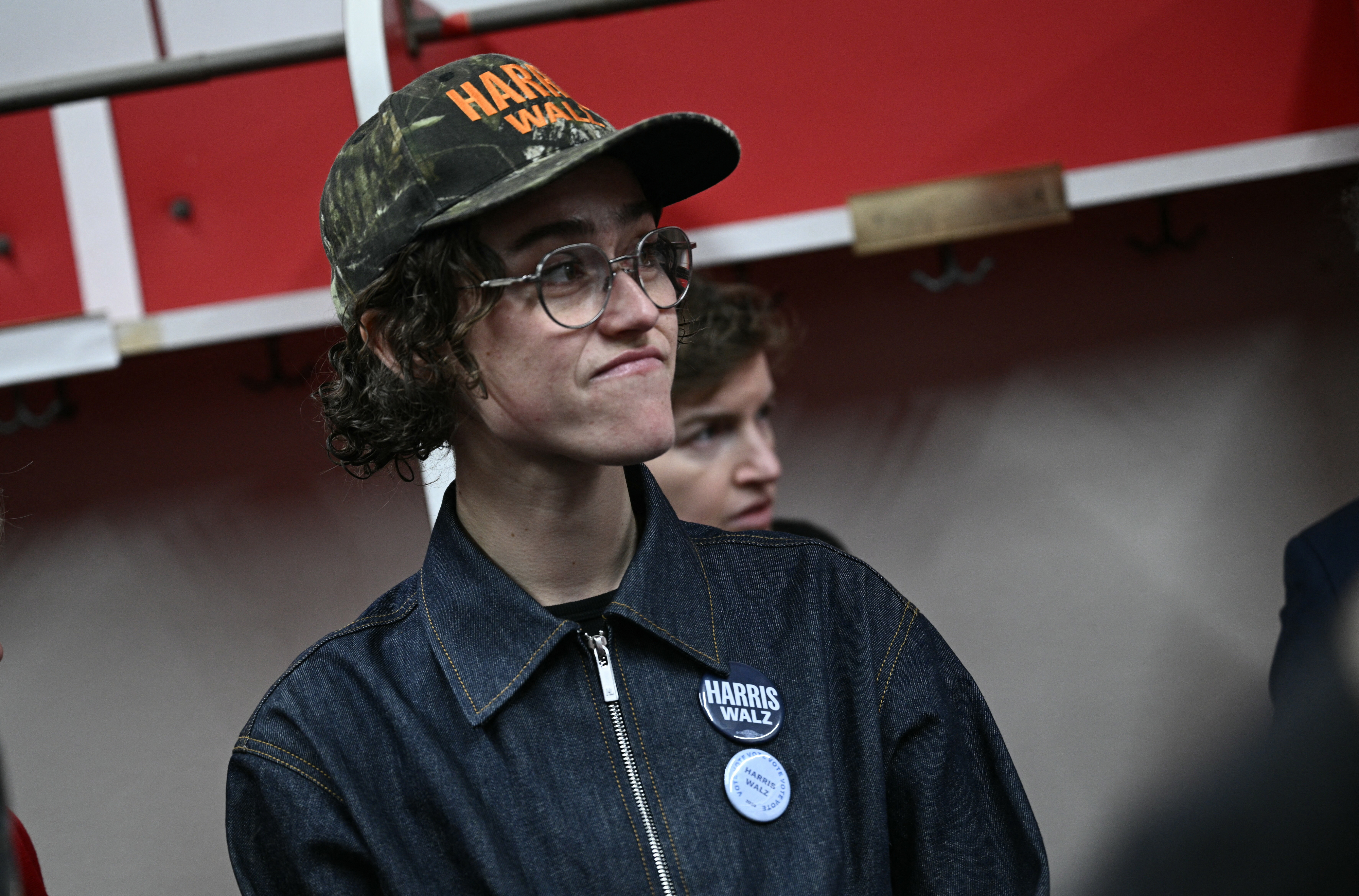 Person wearing a cap and glasses in a denim jacket with a pin, standing in an indoor setting