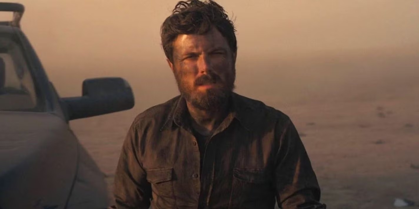 A rugged man with a beard stands in a dusty environment next to a vehicle, looking serious