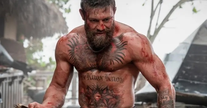 A muscular man with tattoos and a beard appears in a gritty, intense scene, with blood and dirt on his body, suggesting an action movie setting