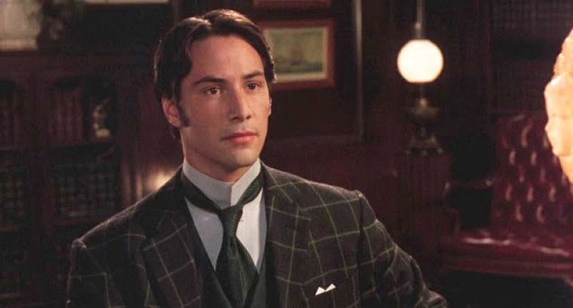 Actor in a plaid suit with a tie stands in a vintage, warmly lit room with framed artwork and a globe lamp