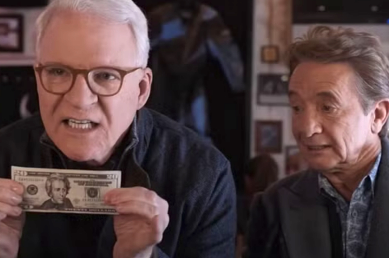 Two men in a room; one is holding a $20 bill and speaking animatedly, while the other observes with a curious expression