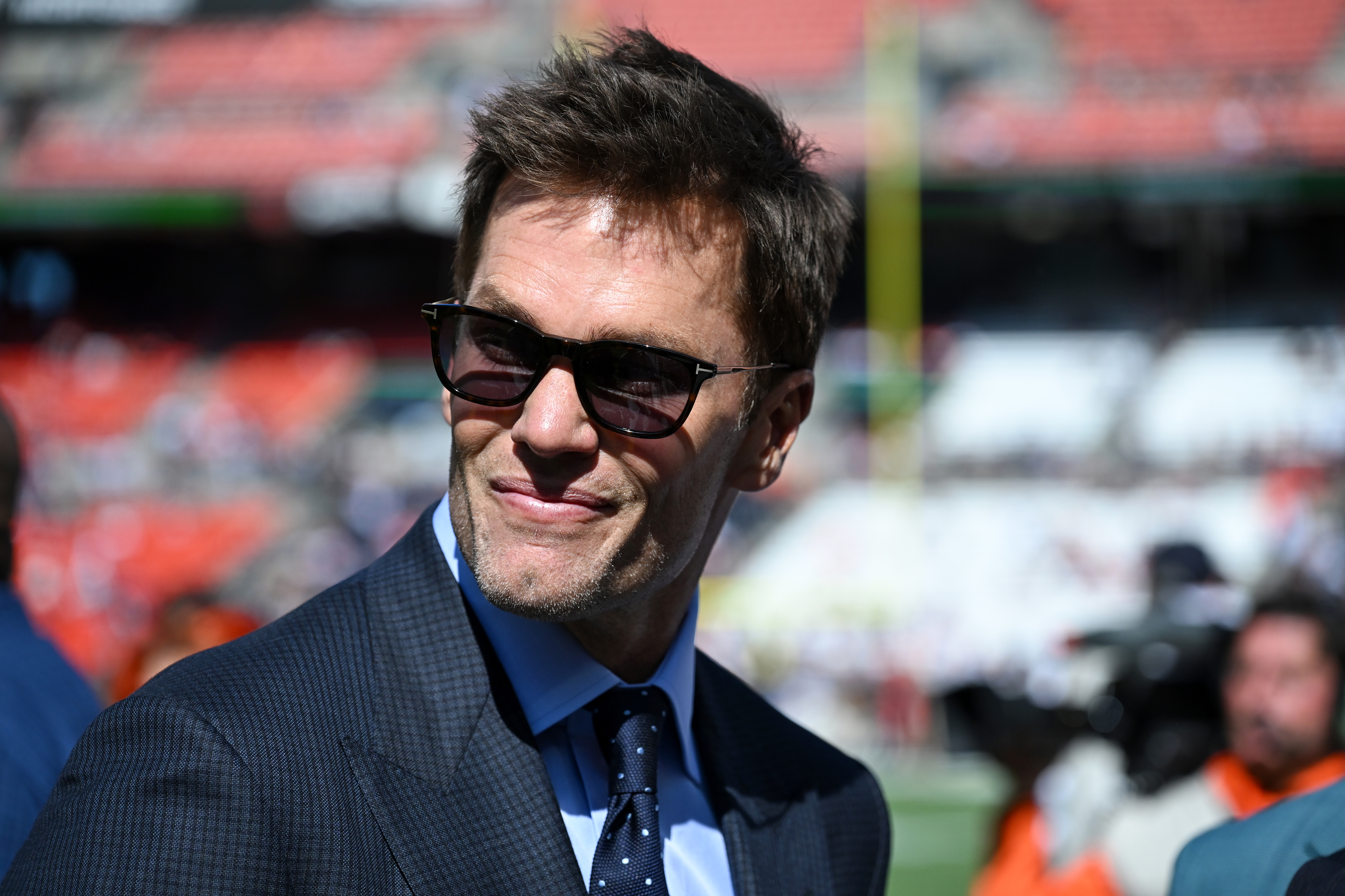 Tom Brady wearing sunglasses and a suit smiles outdoors