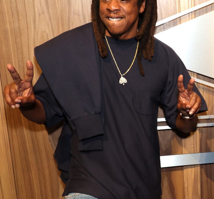 Person wearing casual jeans and a dark shirt, smiling and making peace signs with both hands. They have long hair styled in locs