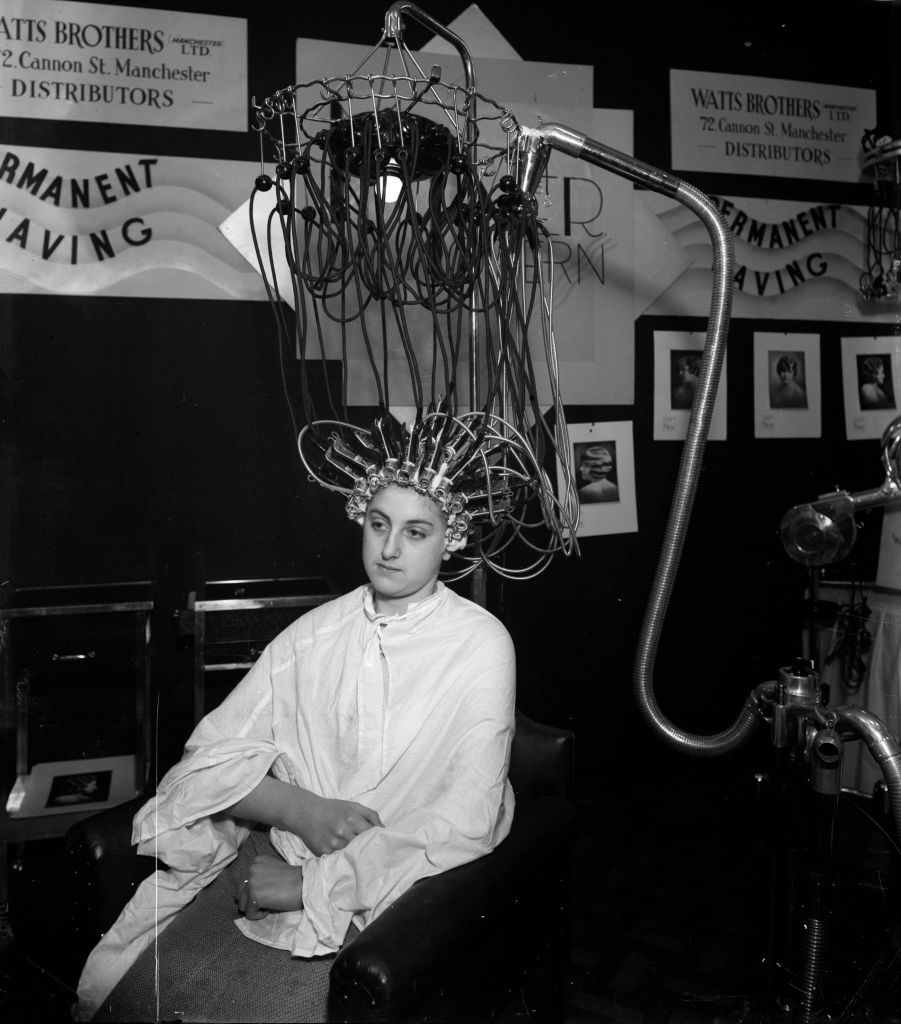 A person sits with an early 20th-century hair perm machine on their head, surrounded by wires, wearing a salon cape