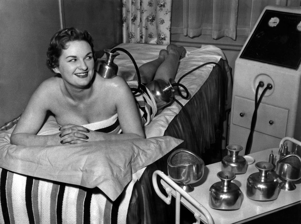 A woman smiles while receiving a vintage beauty treatment involving metal equipment in a spa-like setting