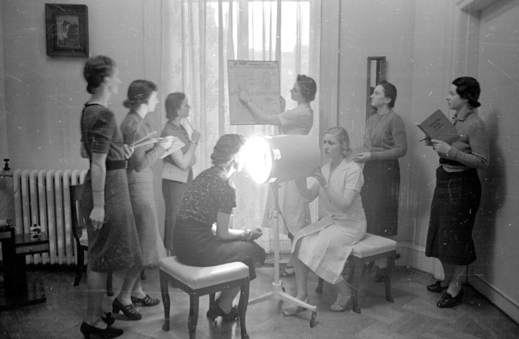 A group of women in 1930s attire stand and sit around a bright spotlight, interacting and reading papers, in a room with a window and radiator