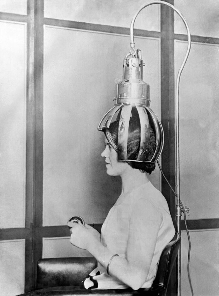 A woman sits with a vintage hair drying machine on her head, holding a control device, in a salon setting