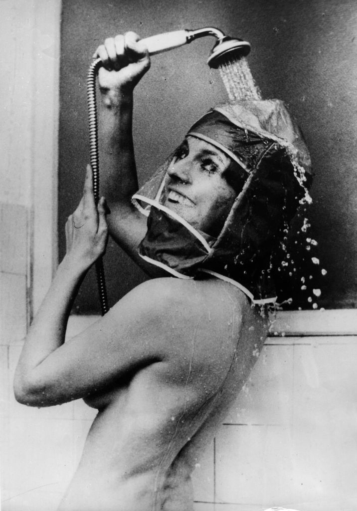 Person using a handheld showerhead while wearing a protective head cover that leaves the face visible, deflecting water away from their face