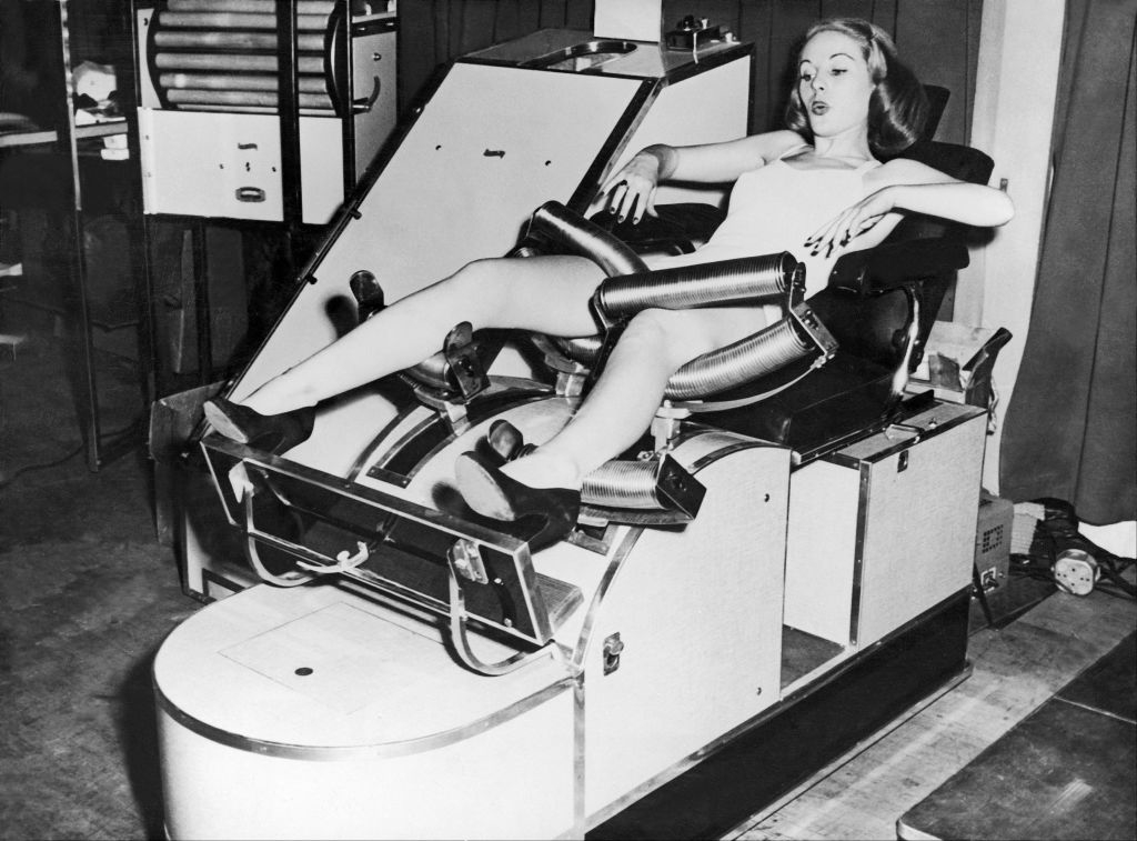 A woman in a swimsuit sits in a vintage exercise machine with leg and arm supports, demonstrating its use